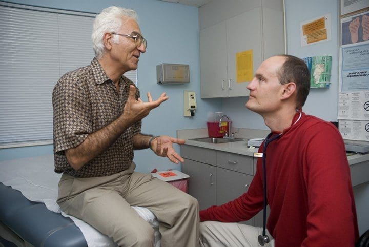 An older man sits on an exam table talking to a healthcare professional in a red shirt with a stethoscope, who is kneeling and listening attentively in a medical office.