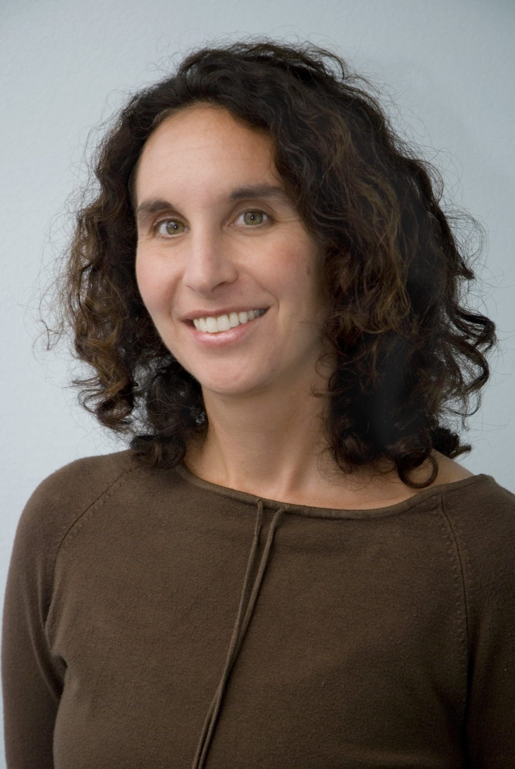 A woman with curly brown hair and light skin smiles at the camera. She is wearing a brown long-sleeve top and is posed in front of a plain light-colored background.