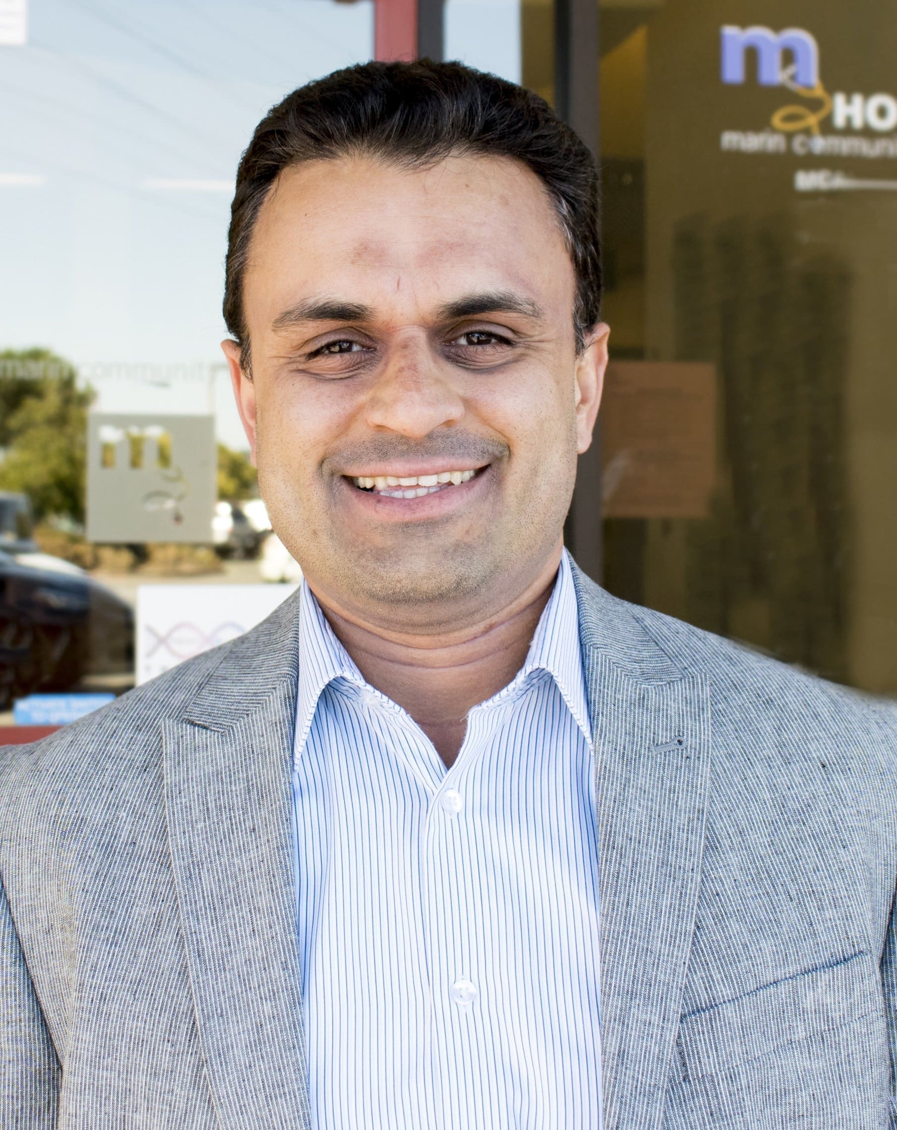 A man wearing a light gray blazer and a striped shirt stands outside a building, smiling at the camera. The background shows glass doors and partial signage with letters and symbols.