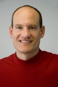 A man with short hair, wearing a red turtleneck shirt, smiles at the camera against a plain, light-colored background.