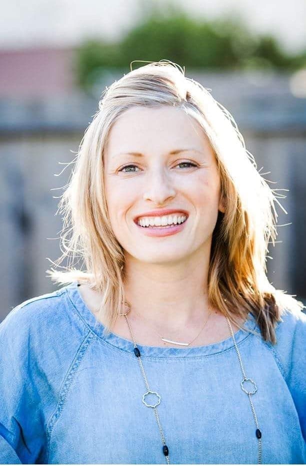 A woman with blonde hair and a bright smile stands outdoors wearing a light blue top and a necklace, with sunlight illuminating her from behind.