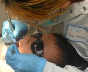 A dentist wearing gloves and a lab coat examines a patients teeth. The patient is lying down and wearing protective sunglasses while the dentist uses dental tools.