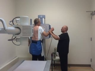 A patient wearing a lead apron stands for a chest X-ray while a technician assists, adjusting the machine in a medical examination room.