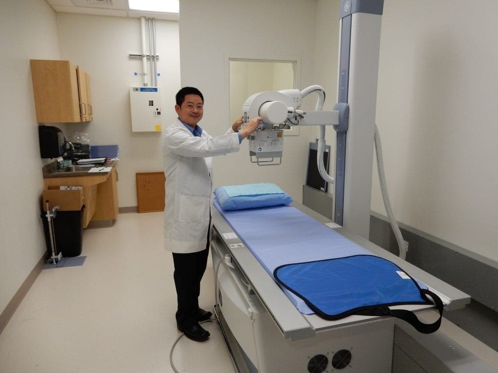 A doctor in a white lab coat stands beside an X-ray machine in a medical examination room, adjusting the equipment above a patient table with a blue pillow and protective apron. A doctor in a white lab coat stands beside an X-ray machine in a medical examination room, adjusting the equipment above a patient table with a blue pillow and protective apron.