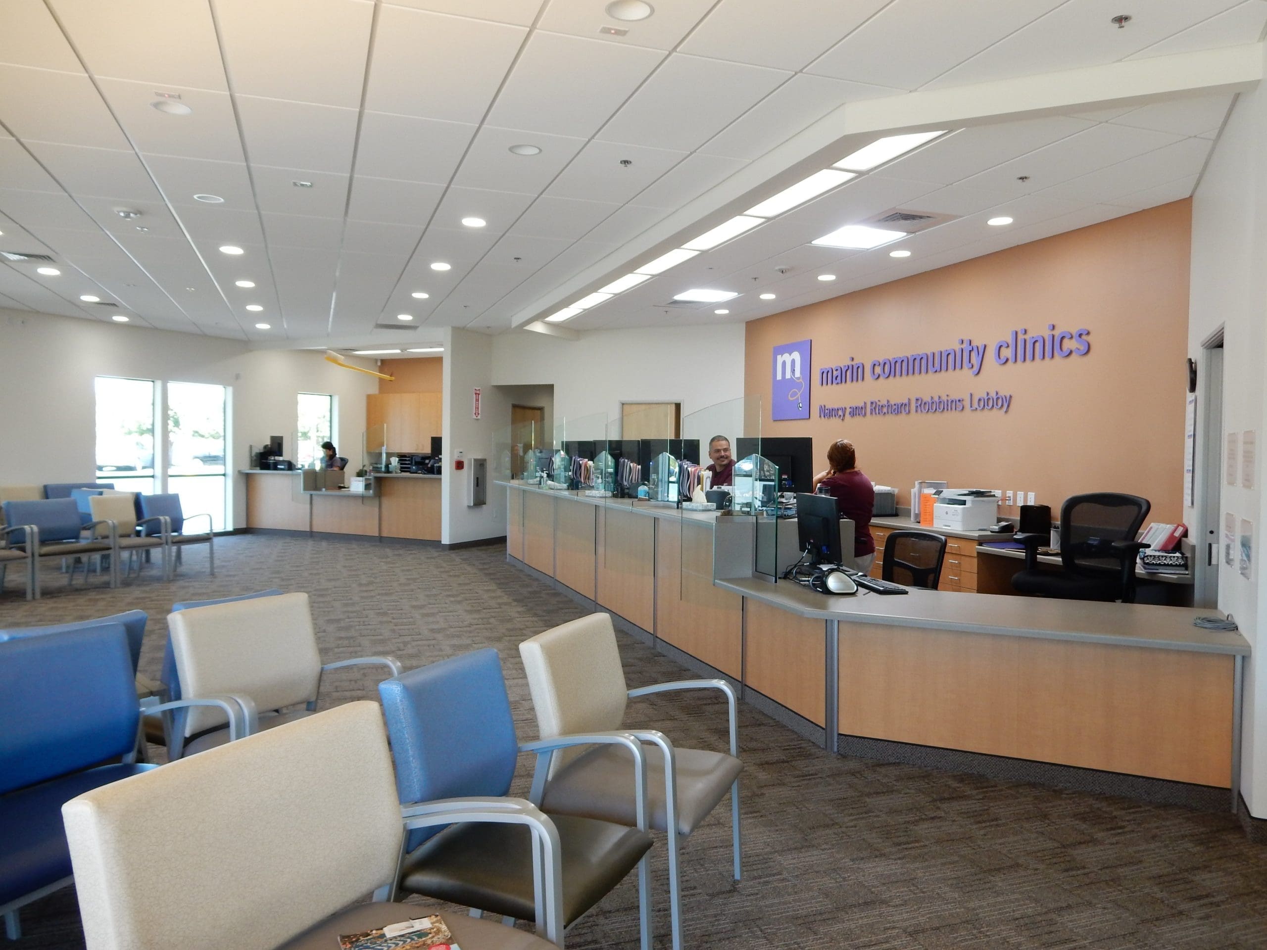 A modern medical clinic waiting area with rows of empty chairs and a front desk where staff assist patients. The wall behind the desk has a sign reading Marin Community Clinics.