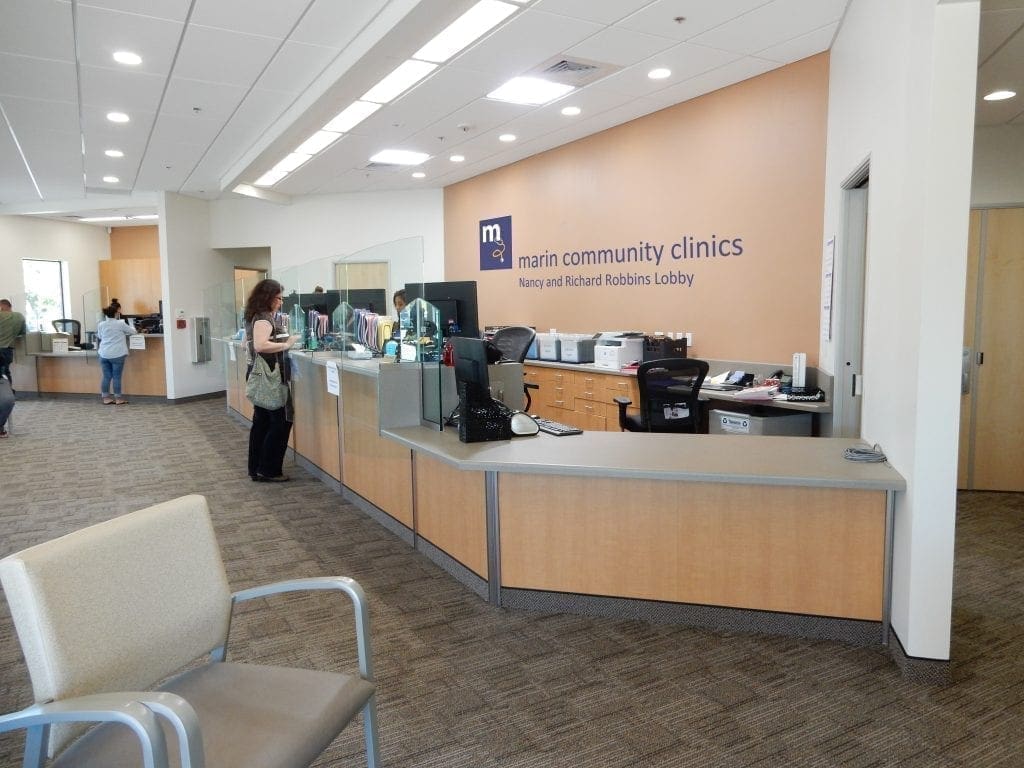 A spacious clinic reception area with a curved desk, staff assisting visitors, and a sign reading “marin community clinics” on the wall. Chairs are arranged in the waiting area, and the space is well-lit and clean. A spacious clinic reception area with a curved desk, staff assisting visitors, and a sign reading “marin community clinics” on the wall. Chairs are arranged in the waiting area, and the space is well-lit and clean.
