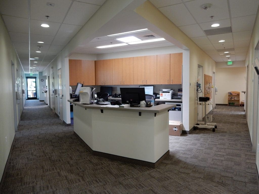 A medical office hallway with a central reception desk, computers, filing trays, and cabinets. The area is clean and organized, with doors leading to exam rooms on both sides and bright overhead lighting. A medical office hallway with a central reception desk, computers, filing trays, and cabinets. The area is clean and organized, with doors leading to exam rooms on both sides and bright overhead lighting.