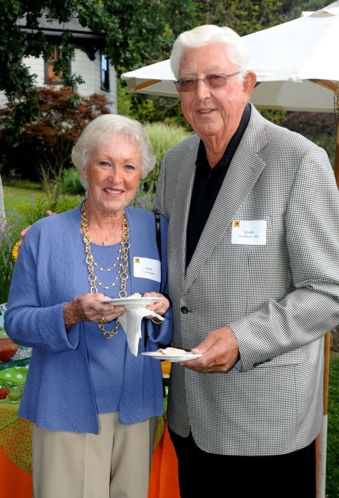 An elderly woman and man stand side by side outdoors at an event, both smiling and holding plates of food. The woman wears a blue jacket, and the man wears glasses and a plaid blazer. They have name tags and are under a white umbrella.