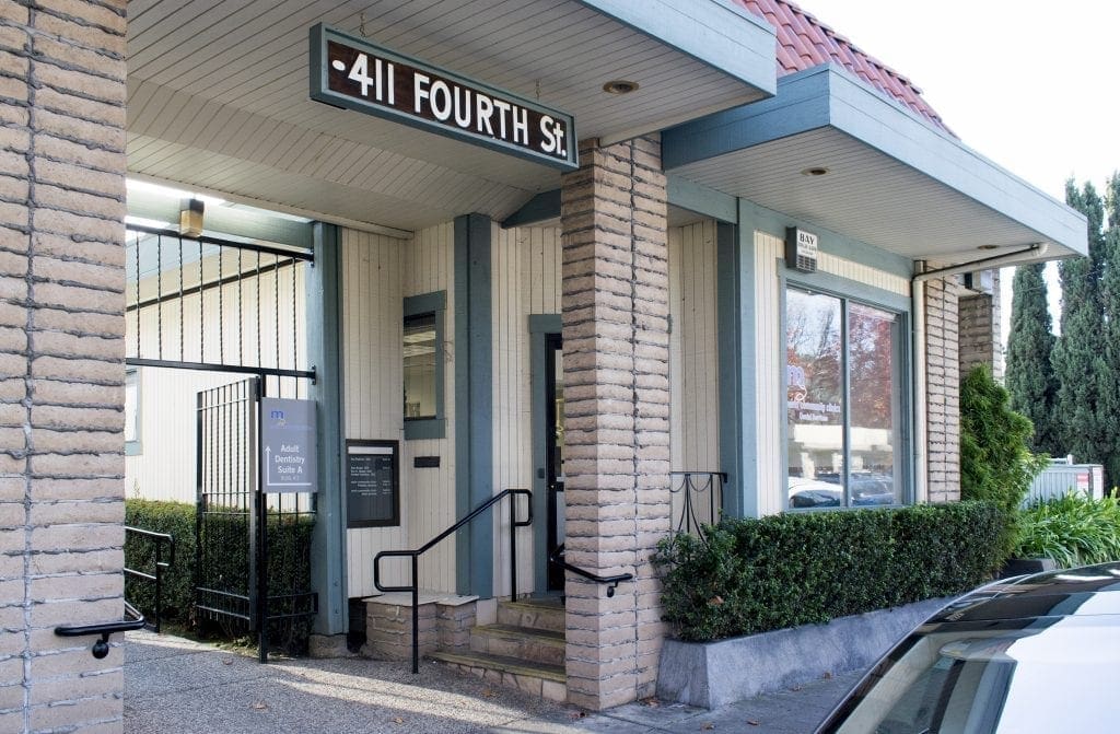 A building entrance at 411 Fourth St. features light brick columns, beige paneling, a black metal gate, steps with railings, and a sign displaying the address above the doorway. Shrubs line the front walkway.