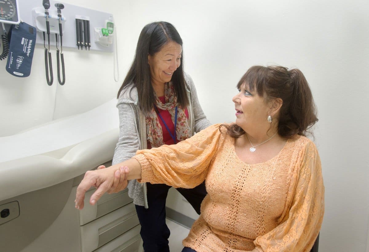 A healthcare worker smiles while examining the arm of a seated woman in a medical exam room. Medical equipment hangs on the wall behind them, and both women appear engaged and comfortable.