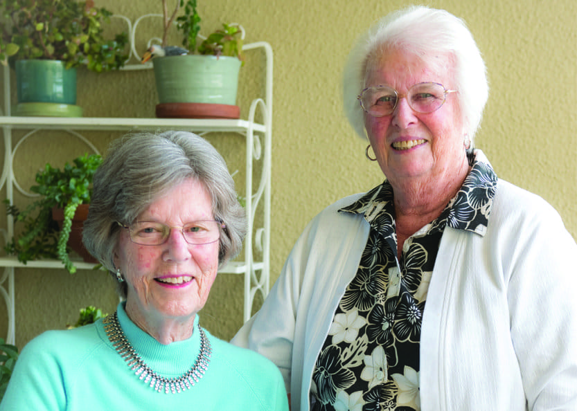 Two elderly women smiling, standing side by side indoors. Both wear glasses; one has on a mint green sweater and necklace, the other wears a white cardigan over a floral blouse. Potted plants are visible behind them.