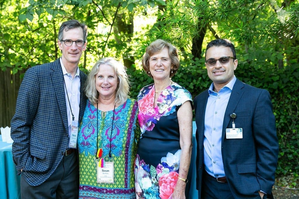 Four adults stand together outdoors, smiling at the camera. Two women are in the middle wearing colorful dresses, while two men in suits stand on either side. Lush greenery and a blue tablecloth are visible in the background.