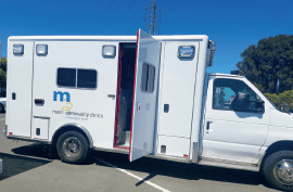 A white mobile medical clinic van with the m logo and main community clinic text on the side is parked outdoors with its side door open under a clear blue sky.