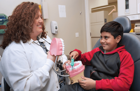 A dentist shows a young boy how to brush teeth using a large dental model and toothbrush. The boy, sitting in a dental chair, smiles and looks at her while practicing.