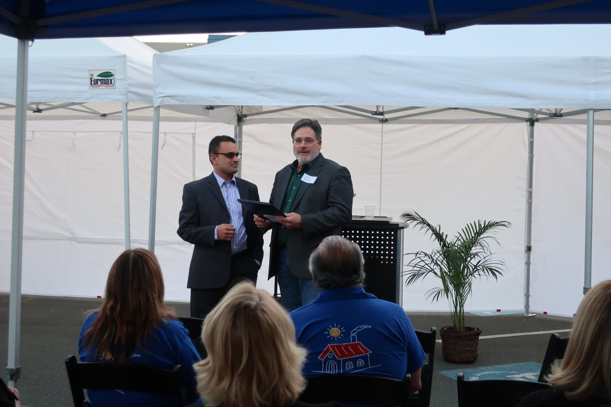Two men in suits stand under a canopy, one holding a framed certificate, speaking to an audience seated in front of them. White tents and a potted plant are visible in the background.