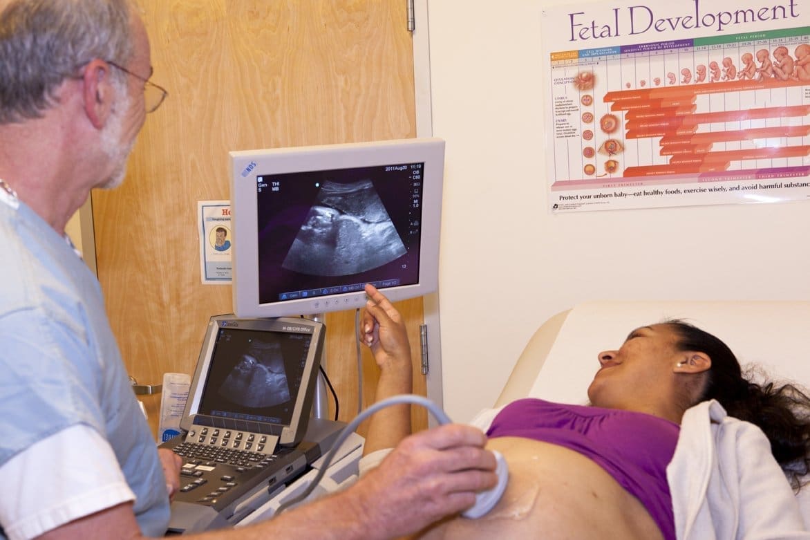 A pregnant woman lies on an exam table while a doctor performs an ultrasound. The doctor points to an ultrasound image on a monitor. A fetal development poster is visible on the wall.