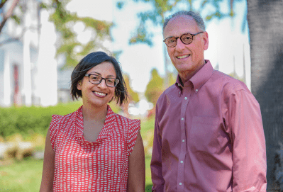 Two people wearing glasses, one in a red patterned blouse and the other in a red button-up shirt, stand smiling outdoors with greenery and trees in the background.