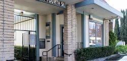 Entrance to a small office building with beige brick columns, a black metal gate, large windows, a railing by concrete steps, and bushes along the front.