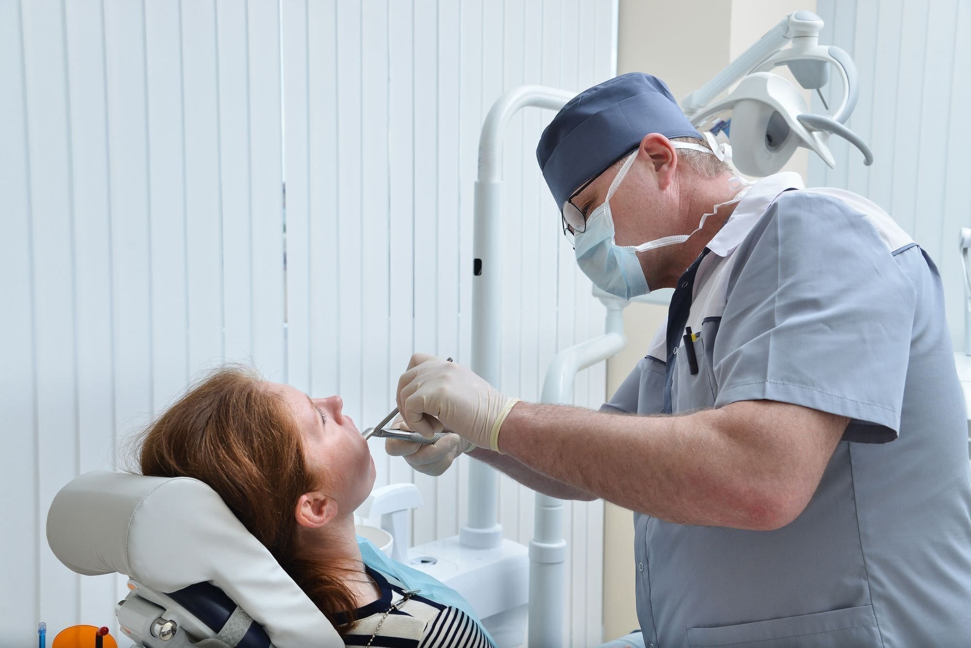 A dentist wearing a mask and gloves examines a female patient’s teeth in a dental clinic, using dental instruments while the patient sits in a reclined chair.