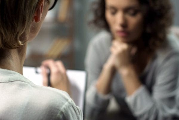 A person with short hair writes on a clipboard while talking to another person with curly hair, who sits with their hands under their chin, appearing thoughtful or concerned.