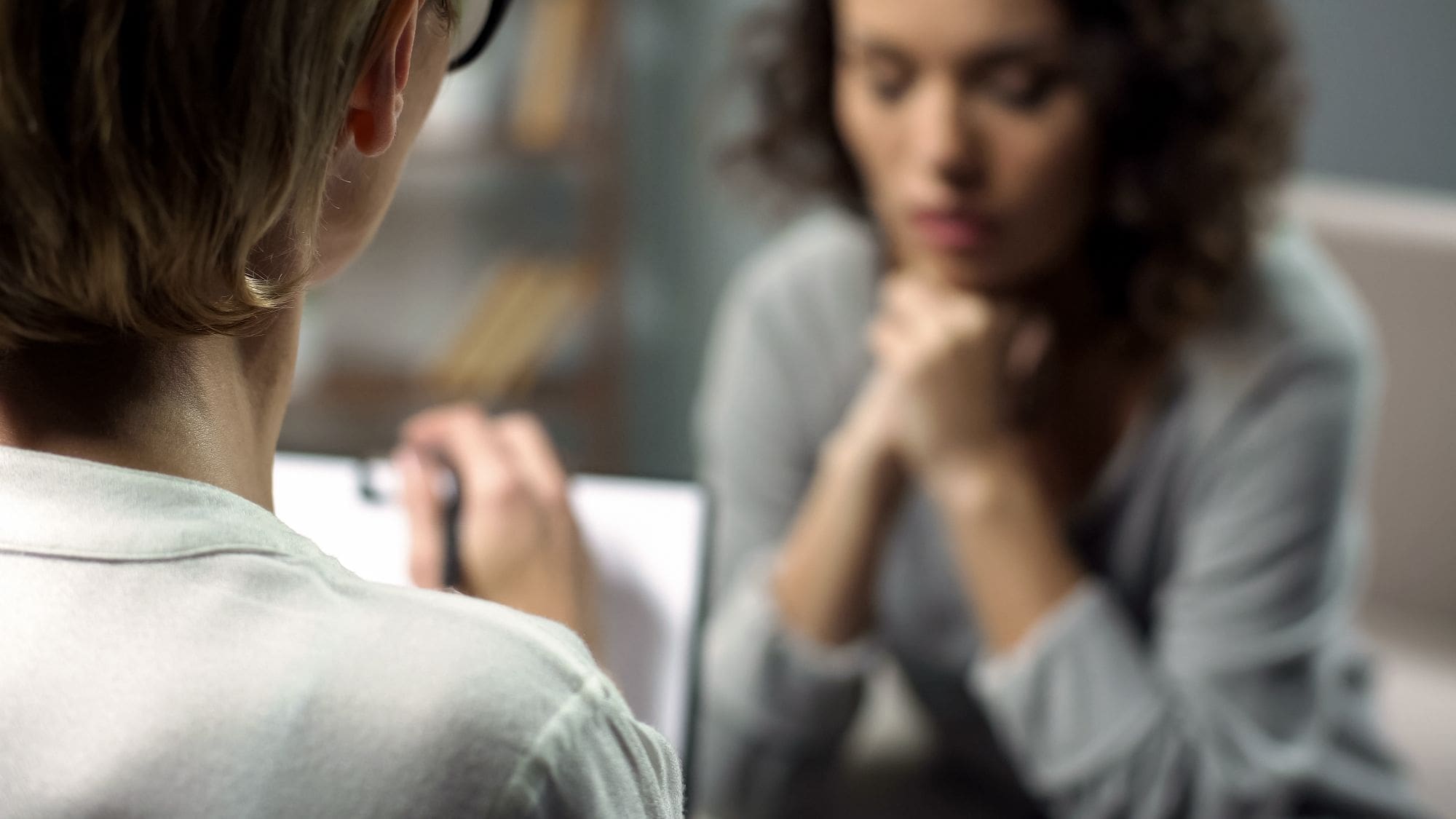A person with short hair writes on a clipboard while talking to another person with curly hair, who sits with their hands under their chin, appearing thoughtful or concerned.