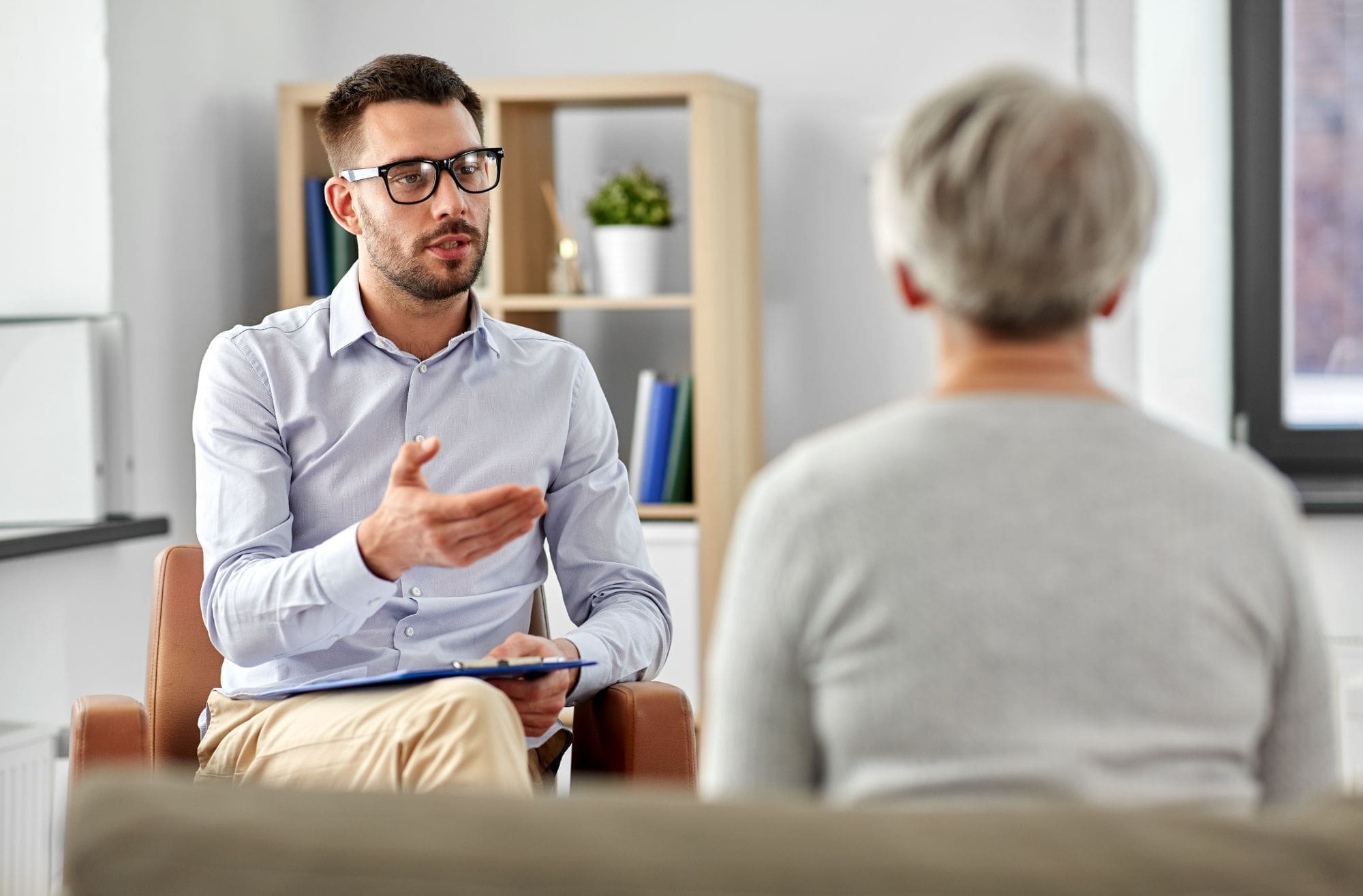 A man with glasses, holding a clipboard, sits and talks to an older person with gray hair in an office setting, suggesting a professional or counseling session. Shelves and a window are visible in the background.