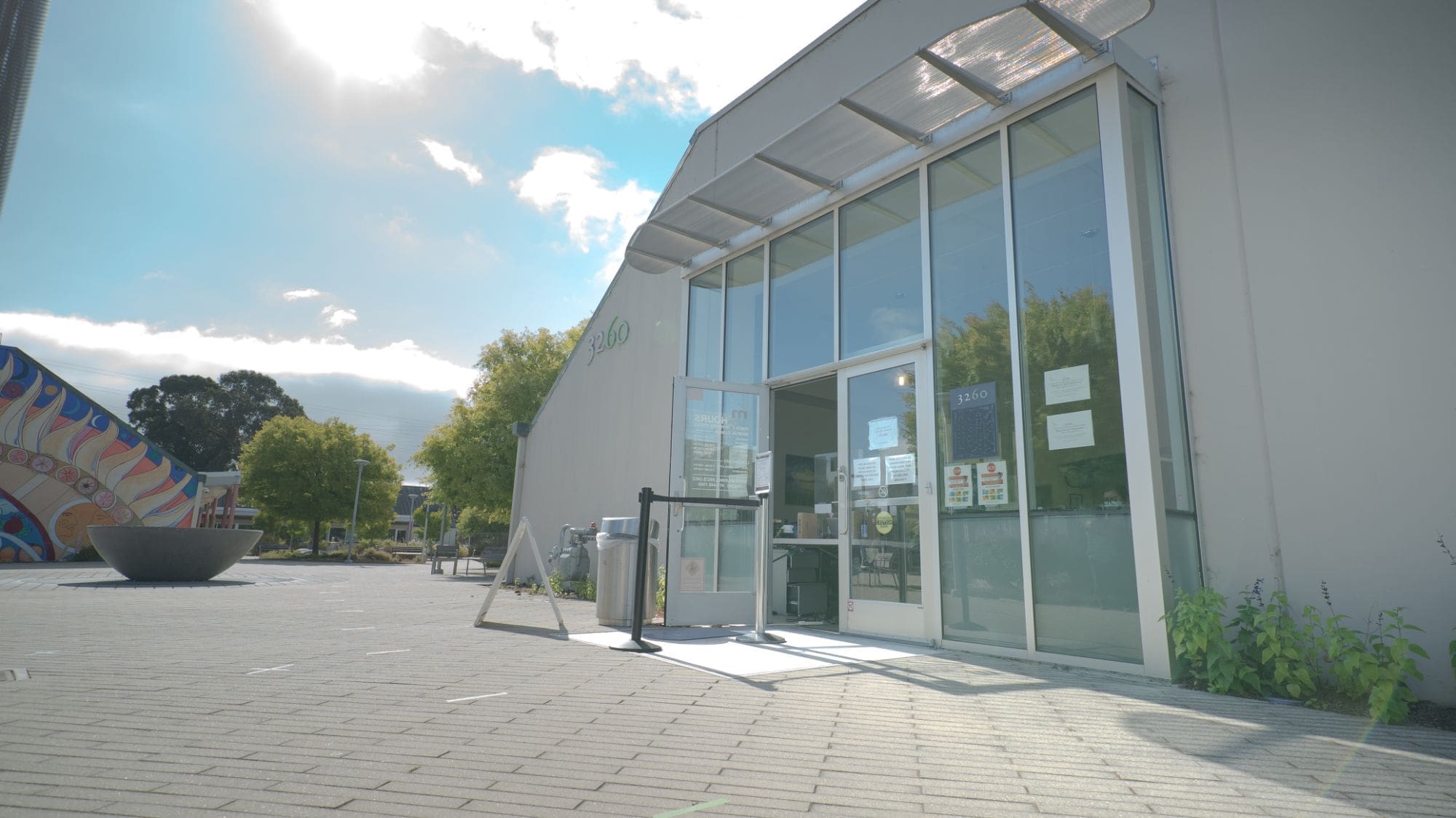 A modern building with large glass doors and windows, featuring signs and a hand sanitizer station at the entrance, is seen on a sunny day with a few clouds in the sky and some trees nearby.