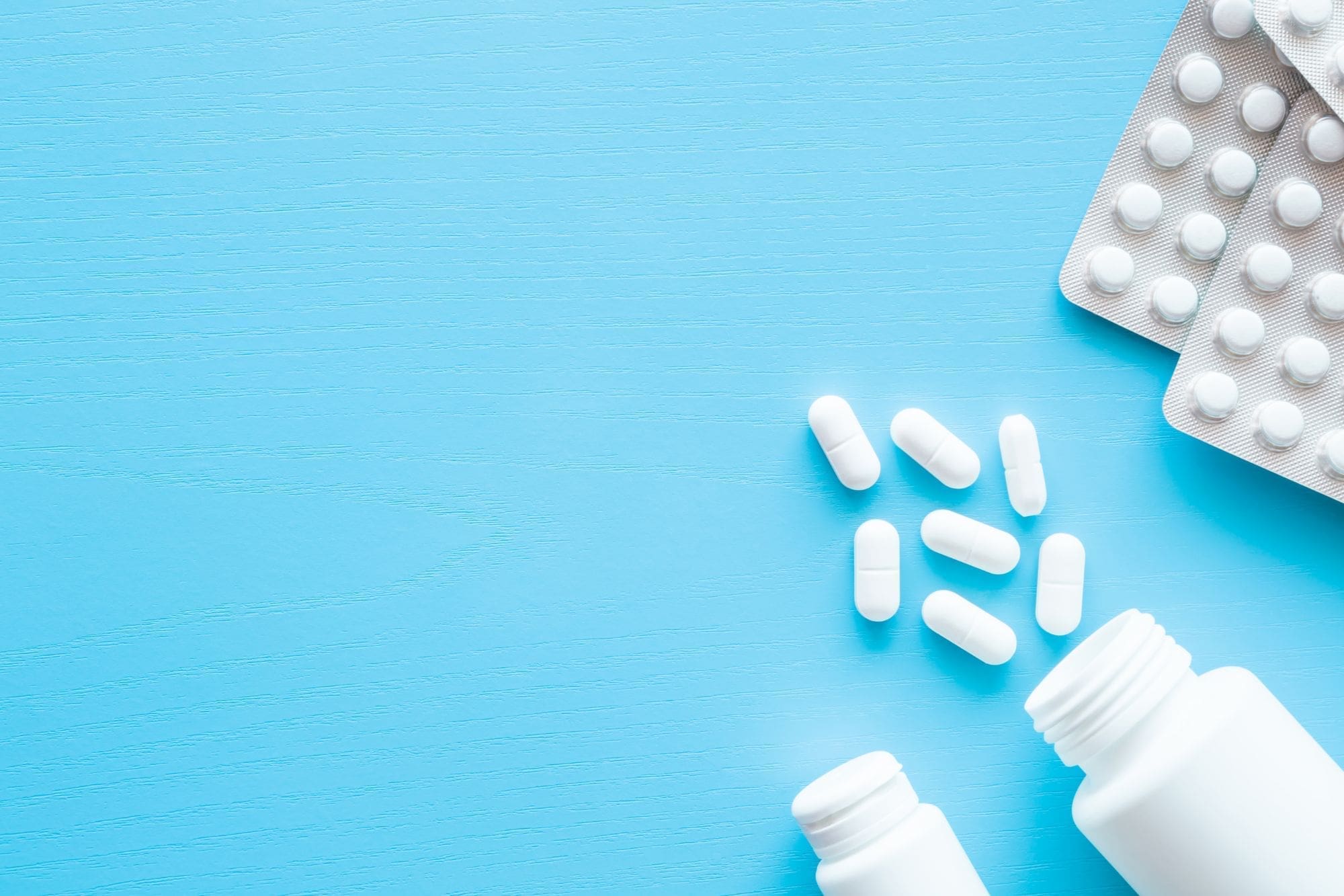 White tablets and capsules scattered on a light blue surface, with two open pill bottles and two blister packs of tablets visible in the top right corner.