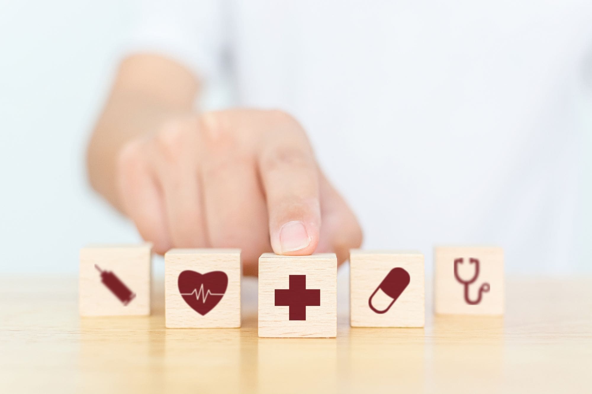 A person points at a wooden block with a red medical cross. Four other blocks with health-related icons—syringe, heart, pill, and stethoscope—are arranged on a wooden surface.