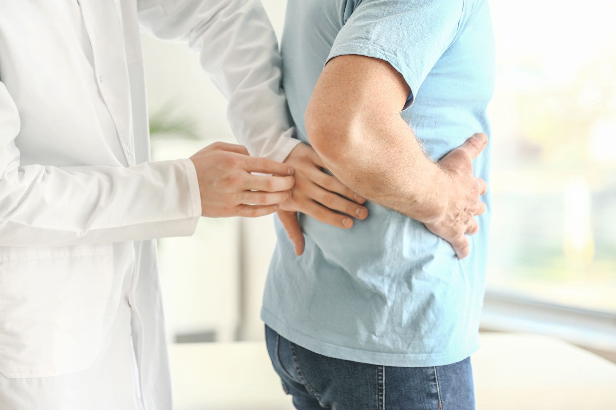 A doctor examines a mans lower back as the man holds his side, suggesting back pain or discomfort. The doctor is wearing a white coat and the patient is in a light blue shirt.