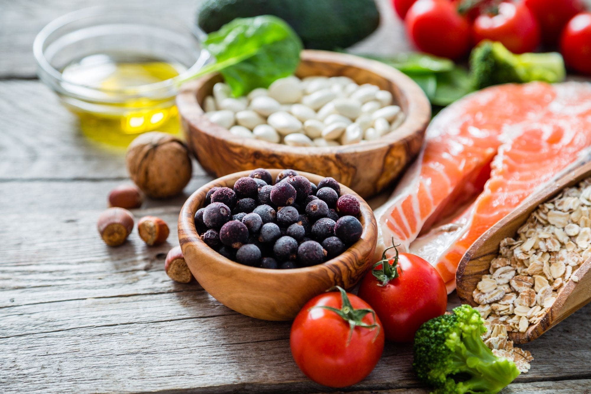 A variety of healthy foods on a wooden table, including salmon, tomatoes, broccoli, oats, blueberries, white beans, walnuts, spinach, avocado, and a small bowl of olive oil.