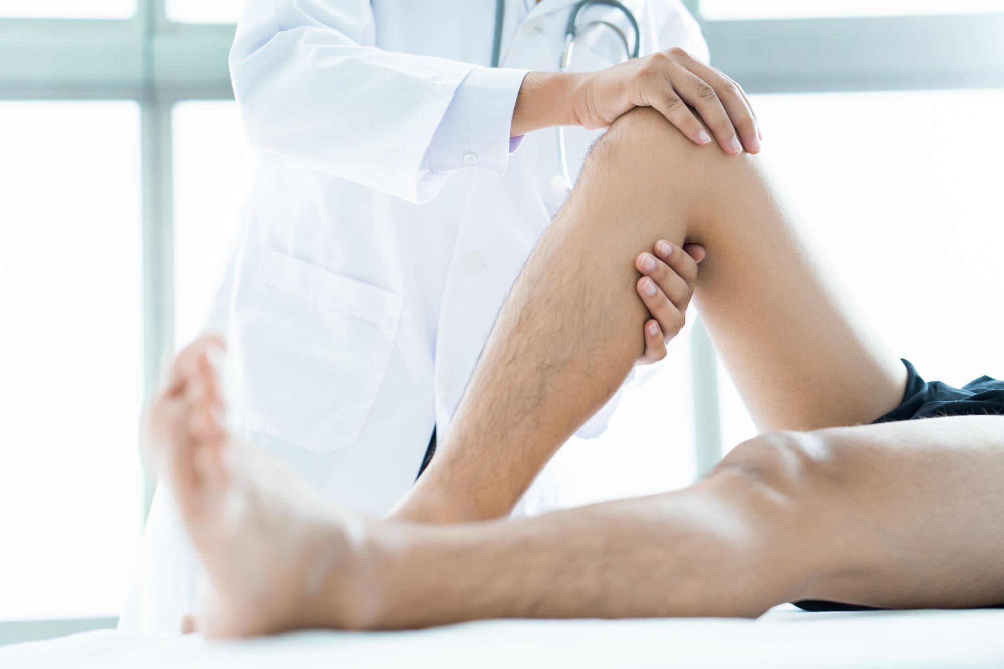 A doctor in a white coat examines a patients bent knee, holding the leg with both hands during a physical assessment in a bright medical office.