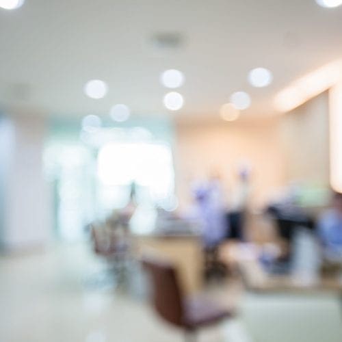 Blurred image of a hospital or clinic reception area with indistinct figures, desks, chairs, and bright lighting, conveying a busy medical or healthcare environment.