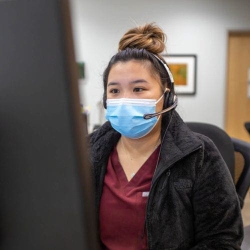 A woman wearing a medical mask and headset sits at a desk, looking at a computer monitor in an office or clinic setting.