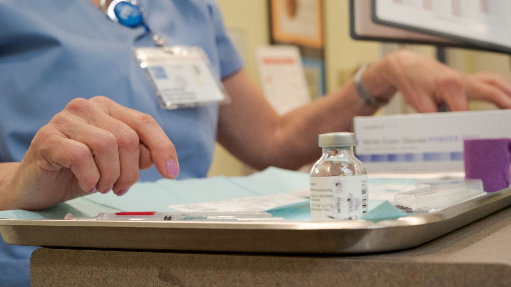 A healthcare worker in scrubs prepares a tray with a vaccine vial, syringes, and alcohol wipes, ready for administration. Only the worker’s hands and torso are visible; their ID badge hangs from their neck.