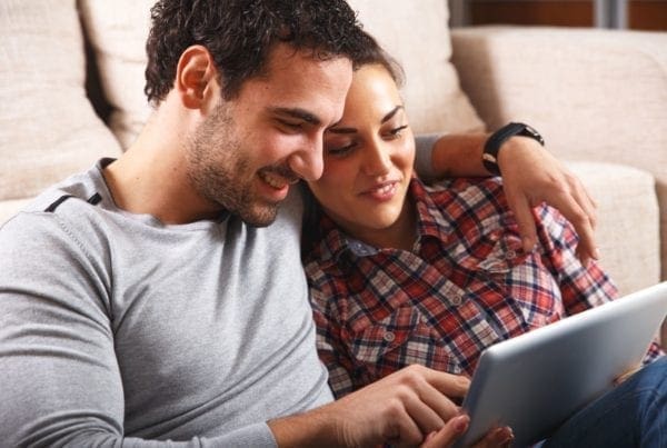 A man and woman sit closely together on a couch, smiling and looking at a tablet screen. The man has his arm around the woman, and both appear relaxed and engaged with the device.