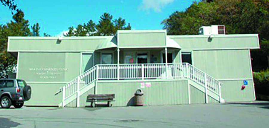 A light green, one-story building with white railings, a ramp, and stairs leading to the entrance. There is a trash can, a bench, and a parked SUV in front. Trees and blue sky are in the background.