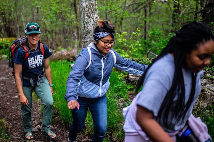 Three people are hiking on a forest trail. Two girls are climbing uphill while an adult with a backpack follows behind. The scene is surrounded by green trees and foliage.