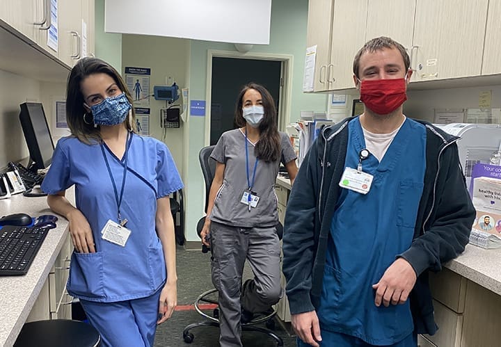 Three healthcare workers wearing scrubs and face masks stand in a medical office, smiling at the camera. Two stand near a counter with computers, while one stands in the background near a door.