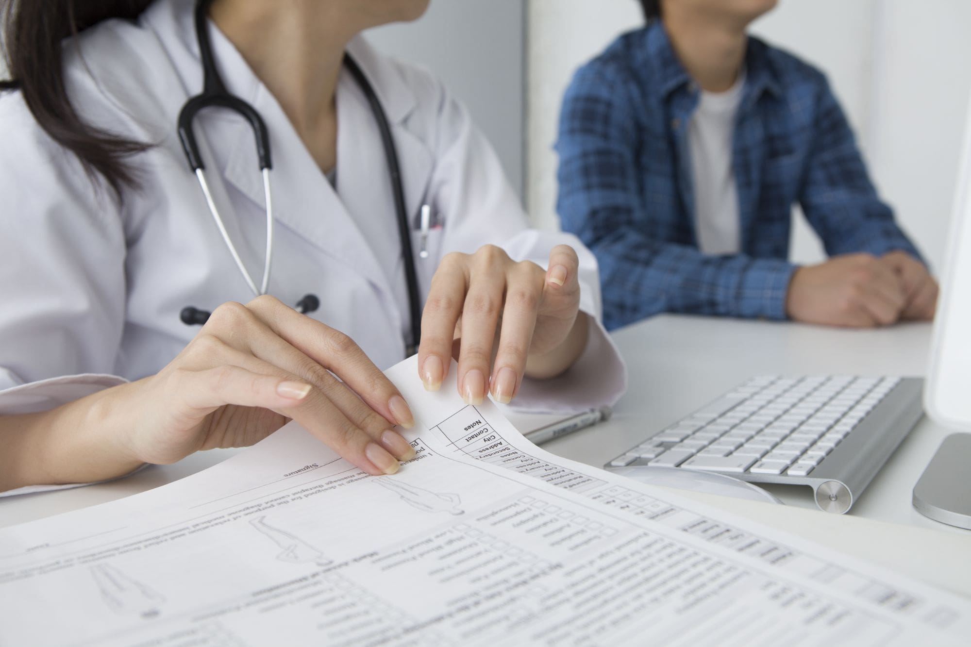 A doctor in a white coat reviews medical documents at a desk with a keyboard, while a patient in casual clothing sits nearby, facing away from the camera.