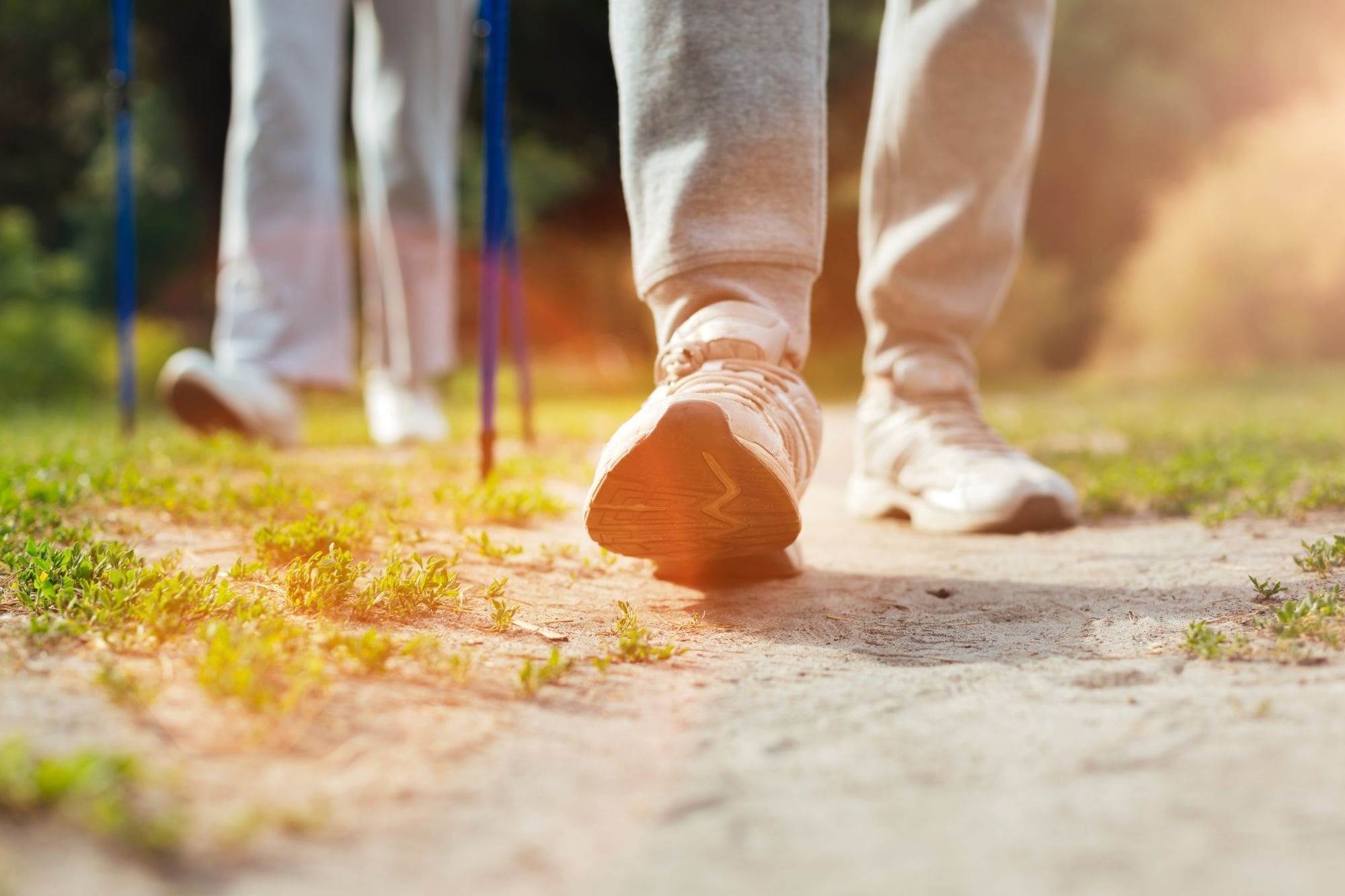 Close-up of two people walking on a sunlit dirt path, wearing light-colored athletic shoes and sweatpants, with one person holding walking poles. Green grass and warm sunlight are visible.