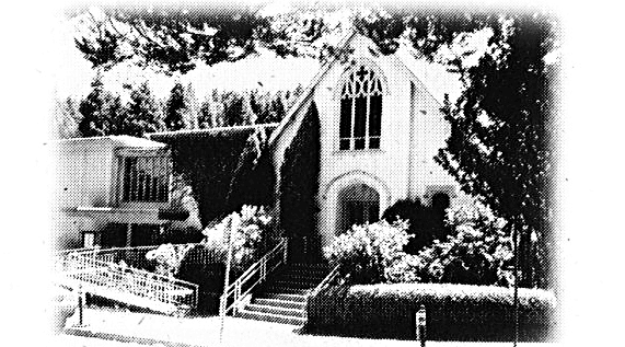 Black-and-white, high-contrast image of a building partially covered by ivy, featuring tall arched windows, a sloped roof, and stairs leading up to the entrance, surrounded by trees and bushes.