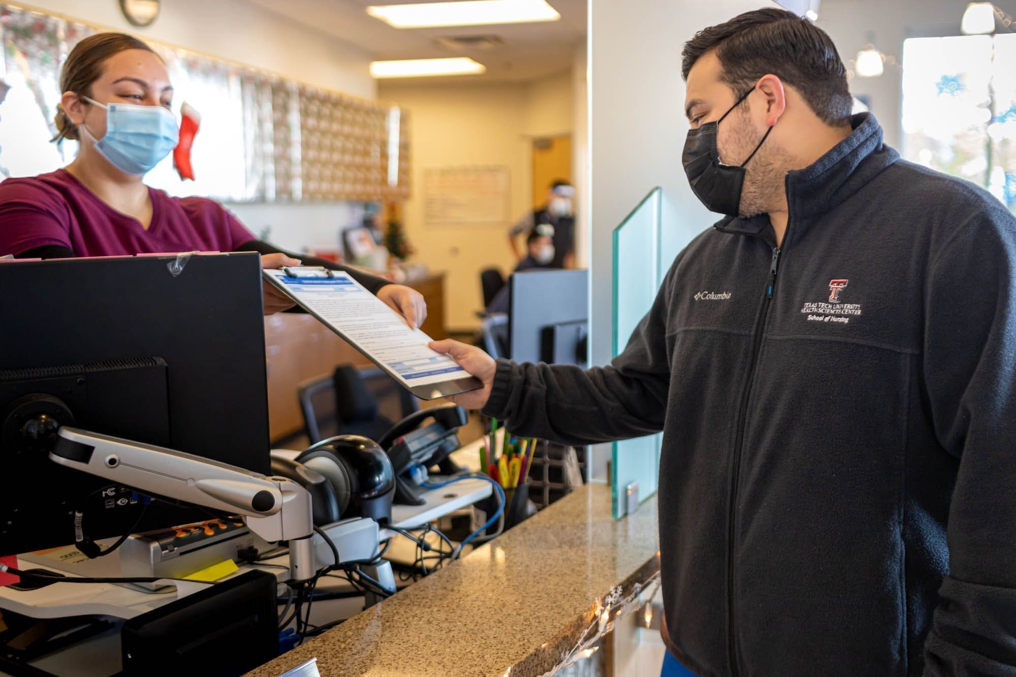 A man wearing a mask hands a clipboard to a masked receptionist at a front desk. The scene appears to be in an office or clinic, with a computer and office supplies on the counter.