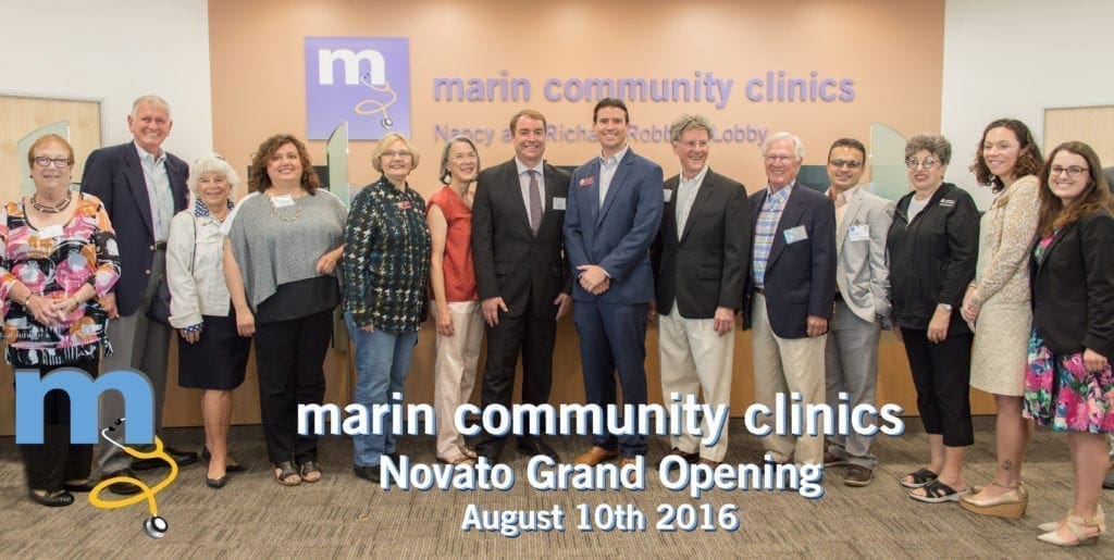A group of people stand smiling in a row inside Marin Community Clinics at the Novato Grand Opening on August 10, 2016. A wall sign with the clinic name and logo is visible behind them.