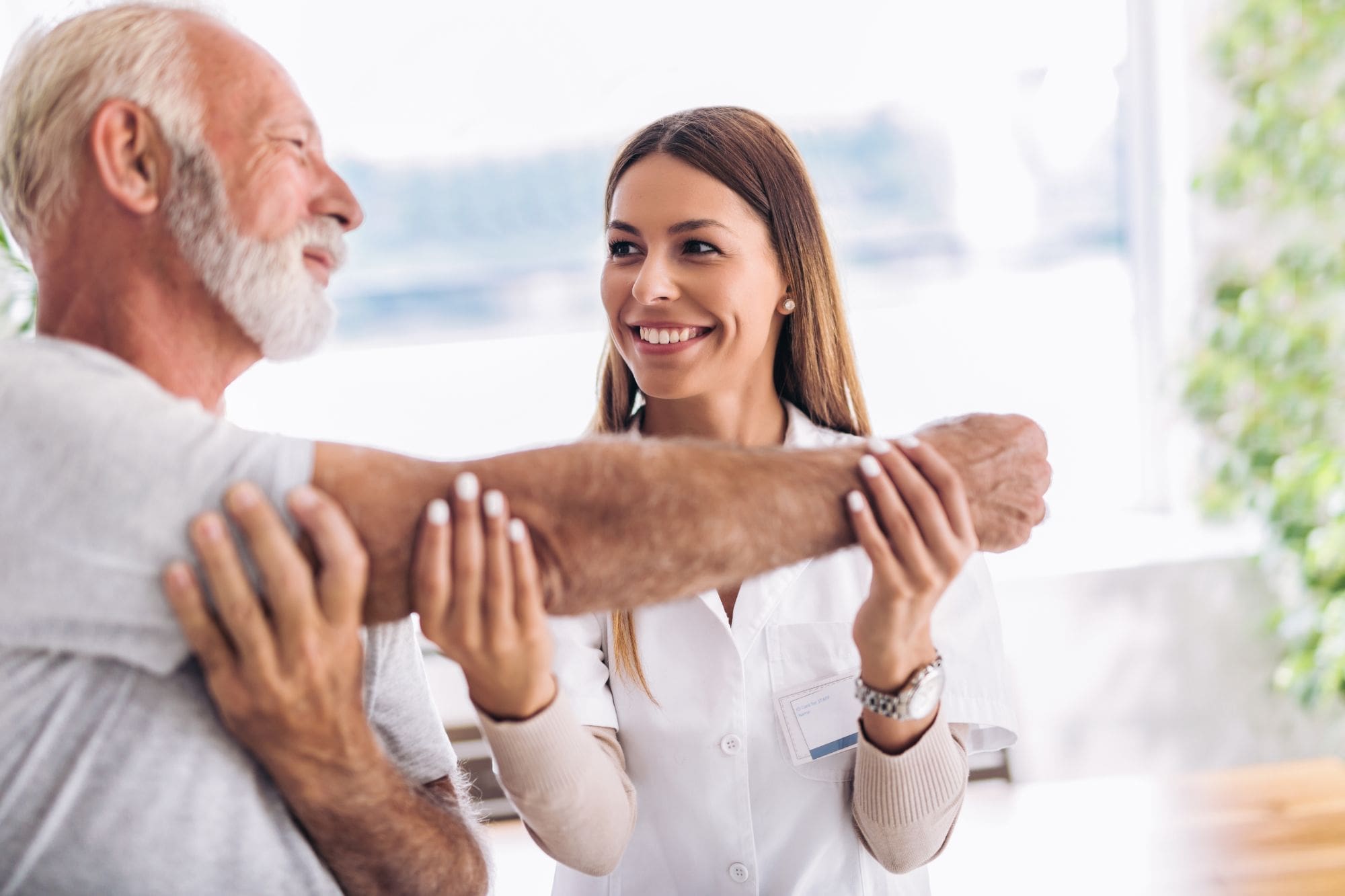 A smiling healthcare professional assists an older man by gently stretching his arm during a physical therapy session in a bright, well-lit room.
