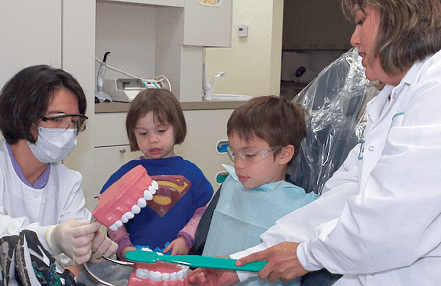 Two dental professionals show a young boy and girl how to brush teeth using a large toothbrush and a model of teeth in a dental office. The children watch attentively, wearing dental bibs.
