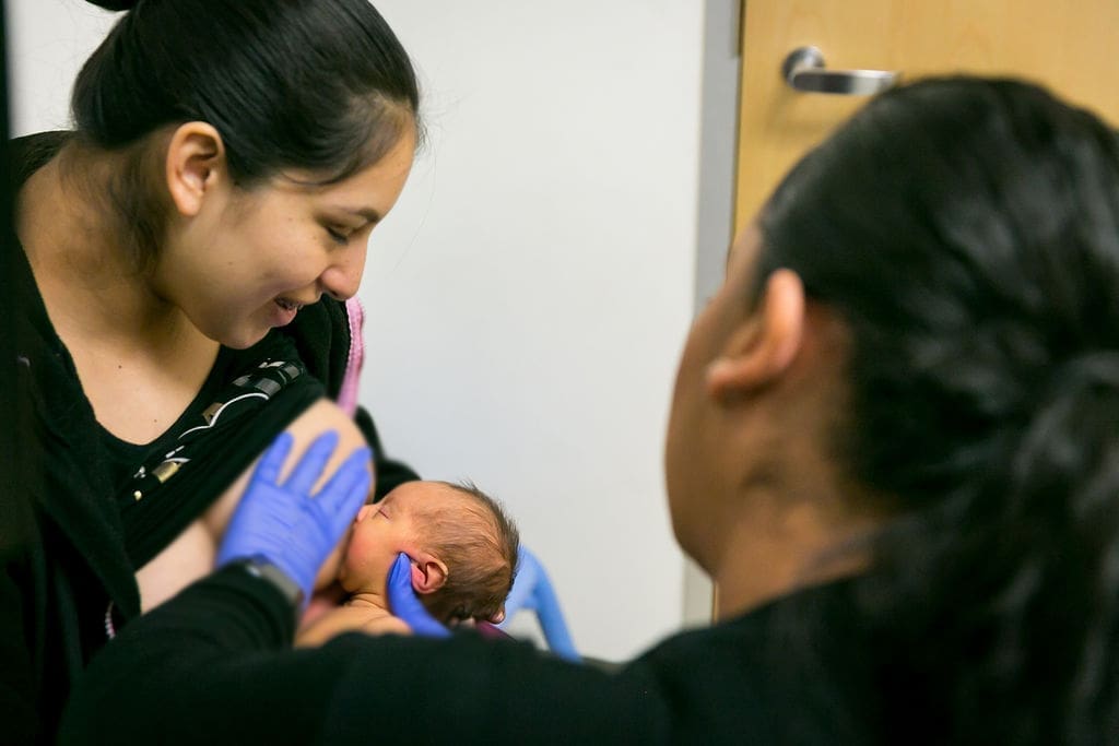 A woman smiles while a healthcare worker wearing blue gloves helps her breastfeed a newborn baby in a clinic room.