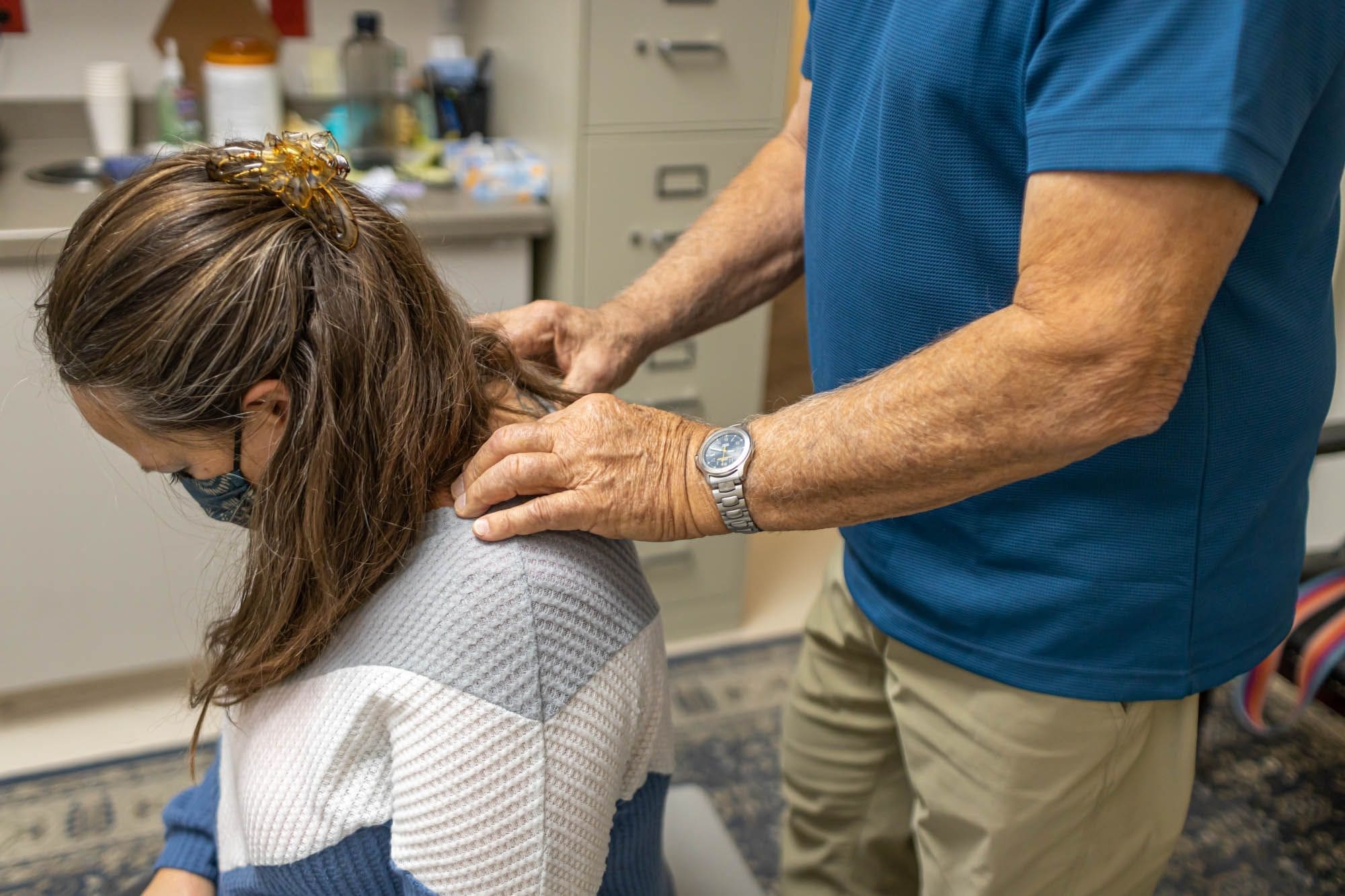 A person in a blue shirt gives a seated woman a neck and upper back massage in an office setting. The woman has a hair clip and is wearing a face mask. Office supplies and cabinets are visible in the background.