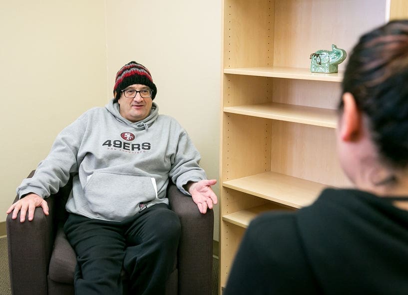 A person wearing a gray 49ers hoodie and a knit hat sits in an armchair, talking to another person whose back is to the camera, in an office with an empty wooden shelf.