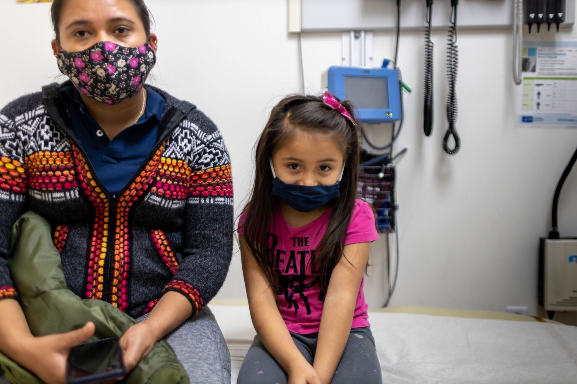 A woman and a young girl wearing face masks sit on an examination table in a medical office. The woman holds a phone, and medical equipment is visible on the wall behind them.